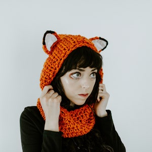 May include: A woman wearing a crocheted fox hat and scarf. The hat has orange and black ears and the scarf is orange.