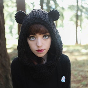 May include: A young person wearing a black hooded hat with bear ears. The hat is crocheted and has a thick, fuzzy texture. The person has long, dark hair and is looking directly at the camera. The background is a blurred forest scene.