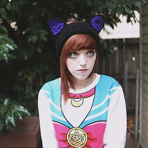 May include: A black knit cat ear beanie with purple accents. The beanie is worn with a white and blue graphic sweatshirt featuring a colorful anime design. The background includes green foliage and a wooden building.