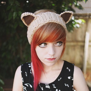 May include: A woman with red and blonde hair is wearing a crocheted headband with cat ears. The headband is beige with brown ears. She is wearing a black and white dress with musical notes printed on it.