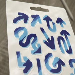 May include: A sheet of stickers featuring various blue arrow designs. The arrows are in shades of light and dark blue, with a pixelated effect. The stickers are on a white backing with a hole for hanging.