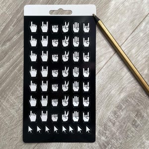 Può includere: Un foglio di adesivi nero con gesti delle mani bianchi e icone del cursore. Una matita color oro è appoggiata su una superficie di legno accanto al foglio. Il foglio ha una parte superiore bianca con un foro per appenderlo.