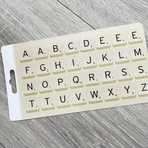 Peut inclure: Une carte rectangulaire avec des tuiles de Scrabble, chacune affichant une lettre et une valeur en points. Les tuiles sont disposées en grille, avec des lettres comme A, B et C visibles. La carte a un trou de suspension et est posée sur une surface en bois gris clair.
