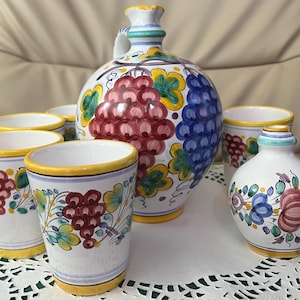 Puede incluir: Un juego de vino de cerámica pintado a mano con una jarra redonda grande con asa, varias tazas pequeñas y una botella pequeña. El juego está decorado con racimos de uvas en rojo y azul, con hojas verdes y detalles amarillos.