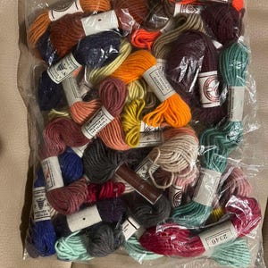 May include: A clear plastic bag filled with various colours of embroidery floss. The floss is in small bundles, with colours including orange, red, green, blue, and yellow. Some bundles have labels with numbers.