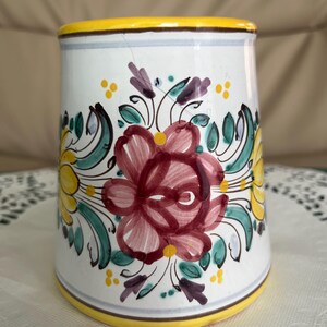 Puede incluir: Jarrón de cerámica blanca con borde y base amarillos, adornado con un diseño floral pintado a mano. Las flores son principalmente rojas y amarillas, con hojas verdes y detalles morados. El jarrón tiene forma cilíndrica.