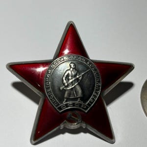 Puede incluir: Una medalla roja en forma de estrella esmaltada con un borde plateado. El centro presenta un soldado con un rifle, rodeado de texto en un diseño circular. La medalla también incluye las letras "CCCP" y un emblema de martillo y hoz.