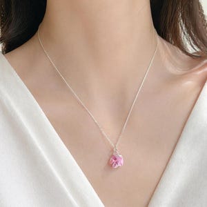 May include: A delicate silver necklace featuring a small, pink flower pendant. The pendant has a textured appearance and is suspended from a fine silver chain. The necklace is worn against a white top.