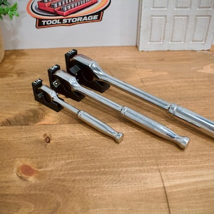May include: Three chrome ratchet wrenches of different sizes are displayed on a wooden surface. Each wrench is held in a black holder. The wrenches are arranged from smallest to largest, with the largest one extending towards the right.