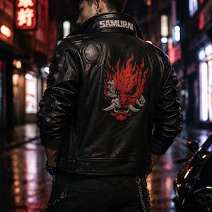 Könnte beinhalten: Eine schwarze Lederjacke mit einer großen rot-schwarzen Grafik einer stilisierten Oni-Maske auf dem Rücken und dem Wort SAMURAI in Weiß auf dem Kragen. Die Jacke verfügt über kreisförmige Details an den Schultern und Ärmeln.