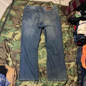 May include: A pair of blue denim jeans shown from the back, featuring two rear pockets with decorative stitching, a waistband with belt loops, and a rectangular leather-like brand patch located on the right side of the waistband above the pocket.