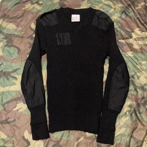 May include: Black V-neck sweater with reinforced patches on the shoulders, elbows, and chest. The long-sleeved sweater has a ribbed knit texture and a small tag near the neckline. The sweater is displayed on a camouflage patterned surface.