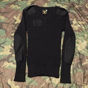 May include: Black ribbed knit sweater with a V-neck, shoulder patches, and elbow patches. The long-sleeved sweater has a small pocket on the chest. The sweater is displayed on a camouflage background.