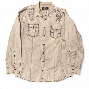 May include: A beige button-up shirt with long sleeves, two chest pockets, and decorative stitching. The shirt has embroidered designs on the shoulders and the brand name "BLACK" on the collar.