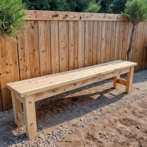 Peut inclure: Banc en bois clair avec une assise rectangulaire et quatre pieds robustes. Le banc est en bois non fini et est placé à l'extérieur près d'une clôture en bois et de petits arbres. Le banc mesure environ 152 cm de long.