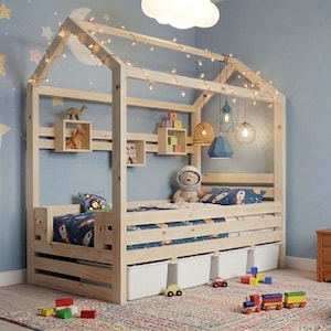 May include: A wooden house-shaped bed frame with a string of lights, a blue patterned comforter, and white storage bins. The room has a blue wall with star and moon decorations, and a rug with toys.