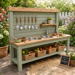 May include: A sage green wooden outdoor play kitchen with a natural wood countertop, two metal sinks, and a faucet. The backsplash features six hanging metal utensils, a small shelf with a potted plant, and three woven baskets filled with produce.