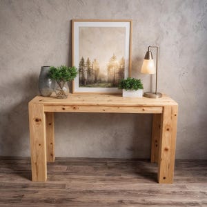 May include: A light-colored wooden console table with a framed landscape print, a small lamp, and decorative vases. The table has a natural wood grain finish and a rectangular shape. The lamp has a metal base and a beige lampshade.