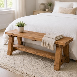 May include: A rectangular wooden bench with a natural brown finish, featuring four sturdy legs and a lower shelf. A small potted green plant sits on the left side of the bench, and a folded beige fringed throw blanket rests on the right side.