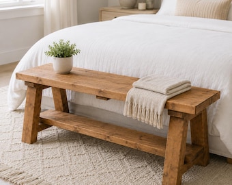 May include: A rectangular wooden bench with a natural brown finish, featuring four sturdy legs and a lower shelf. A small potted green plant sits on the left side of the bench, and a folded beige fringed throw blanket rests on the right side.