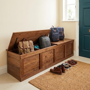 Könnte beinhalten: Eine große Holz-Aufbewahrungsbank mit Klappdeckel, gefüllt mit Rucksäcken und Taschen. Die Bank hat eine warme braune Farbe und eine rechteckige Form. Mehrere Paar Schuhe sind vor der Bank auf einem gewebten Teppich aufgereiht. Die Bank ist etwa 150 cm lang.