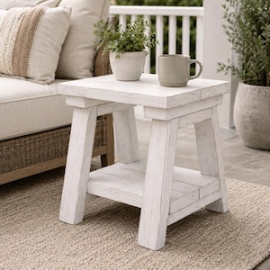 Peut inclure: Une table d'appoint en bois blanc avec une finition vieillie, dotée d'un plateau carré et d'une étagère inférieure. La table possède quatre pieds robustes et est faite de planches épaisses. Une petite plante verte et une tasse grise sont posées dessus.