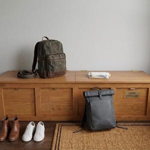 May include: A wooden shoe storage bench with a dark green and brown backpack, a gray backpack, and two pairs of shoes. The bench has four drawers and a brass "SHOE STORAGE" sign.