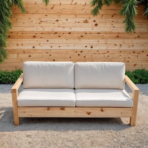 以下が含まれることがあります： 2つのクリーム色のクッションが付いた、明るい色の木製屋外用ラブシート。 ラブシートは、天然木のフレームを使用したシンプルでモダンなデザインです。 背景には木のフェンスと緑があり、パティオや庭の設定を示唆しています。