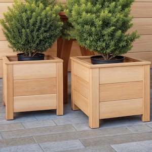 Könnte beinhalten: Zwei quadratische Pflanzgefäße aus hellem Holz, jedes mit einem dreiteiligen Design, die grüne Pflanzen in schwarzen Töpfen enthalten. Die Pflanzgefäße stehen auf kurzen Beinen. Der Hintergrund zeigt eine Holzwand und einen gefliesten Boden. Jedes Pflanzgefäß ist etwa 40 cm breit.