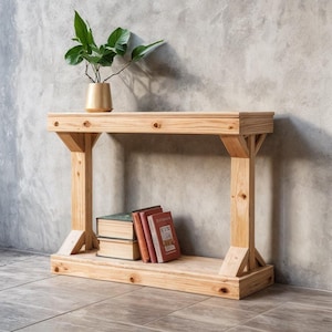 May include: A natural wood console table with a lower shelf, holding several books. A small gold-colored pot with a green plant sits on the tabletop. The table is made of light-colored wood and has a simple, sturdy design.