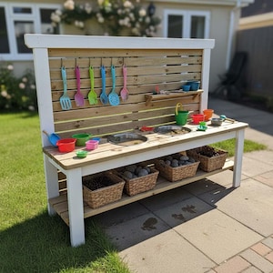 May include: A wooden outdoor play kitchen with a white frame and a natural wood back panel. It features a sink, shelves, and hanging utensils in various colors. Baskets filled with rocks are on the bottom shelf, and colorful toys are on the countertop.