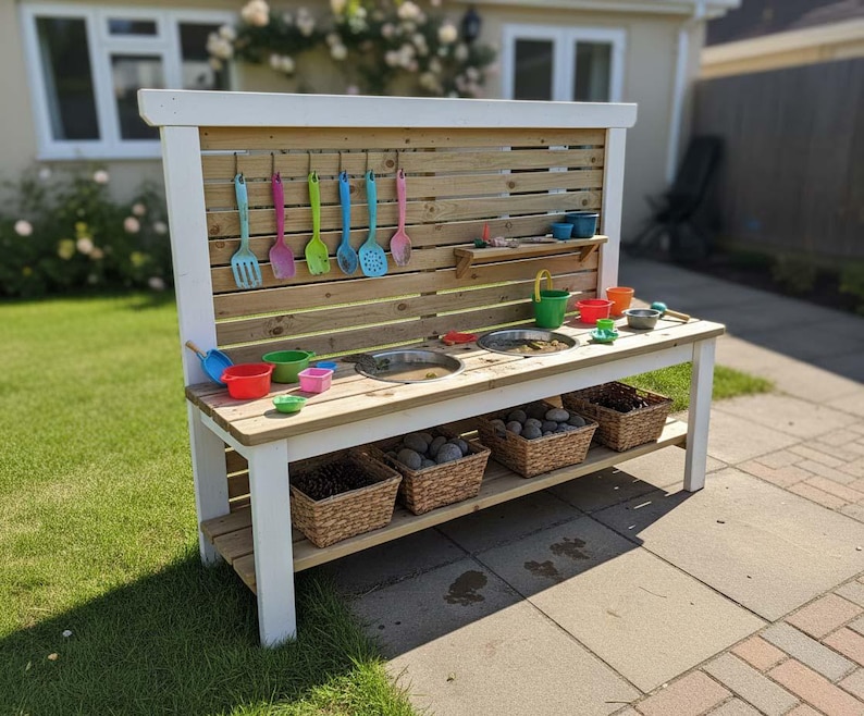 May include: A wooden outdoor play kitchen with a white frame and a natural wood back panel. It features a sink, shelves, and hanging utensils in various colors. Baskets filled with rocks are on the bottom shelf, and colorful toys are on the countertop.