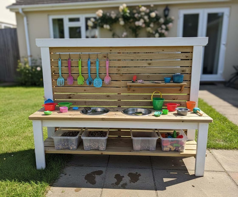 May include: An outdoor wooden play kitchen with a white frame and a natural wood backsplash. It has two stainless steel sinks, colourful utensils, and storage bins. The play kitchen is designed for children.