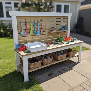 May include: A wooden outdoor play kitchen with a white frame and a natural wood back panel. It features a sink, a wash basin, and various colorful utensils and bowls. Baskets filled with rocks are stored on a lower shelf.