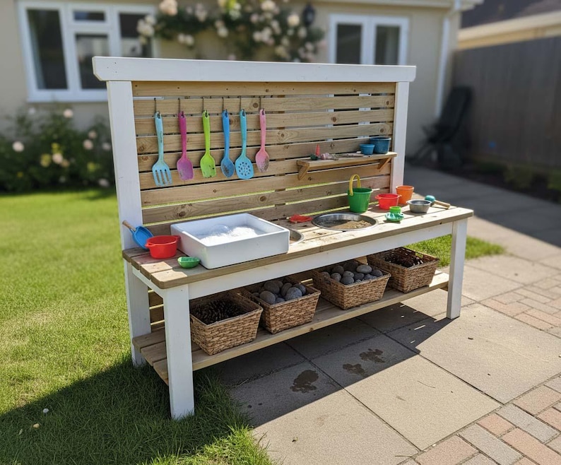 May include: A wooden outdoor play kitchen with a white frame and a natural wood back panel. It features a sink, a wash basin, and various colorful utensils and bowls. Baskets filled with rocks are stored on a lower shelf.