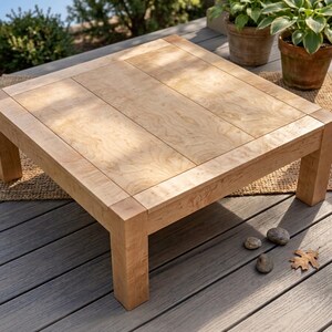May include: Square wooden coffee table with a light wood grain finish. The table has a simple, modern design with a thick frame and legs. The tabletop is made of several planks of wood. The table is approximately 36 inches square.