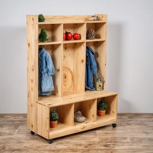 Puede incluir: Un organizador de entrada de madera clara con múltiples cubículos, estantes y un banco. Dos chaquetas vaqueras cuelgan en el interior. Los elementos decorativos incluyen pequeñas plantas, manzanas rojas y una piña. La unidad está sobre ruedas.