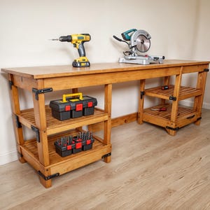 Könnte beinhalten: Eine Werkbank aus Holz mit zwei Regalebenen und schwarzen Metallecken. Ein gelber und schwarzer Akku-Bohrschrauber und eine silberne Gehrungssäge stehen auf der Tischplatte. Zwei schwarze Werkzeugkästen mit roten Akzenten befinden sich auf den Regalen.