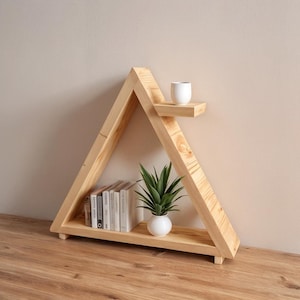 May include: A wooden, triangular-shaped shelf unit with two shelves. The bottom shelf holds a small stack of books and a white ceramic pot with a green plant. A small white cup sits on the top shelf. The unit is made of light-colored wood.