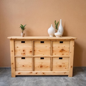 Puede incluir: Una cómoda de madera clara con seis cajones, dispuestos en dos filas de tres. La cómoda tiene un acabado de veta de madera natural. Tres jarrones de cerámica blanca y una pequeña planta en maceta encima.