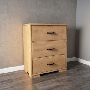 May include: A three-drawer wooden dresser with a light oak finish. The dresser has black handles and sits on a low base. The drawers are evenly spaced and the wood grain is visible.