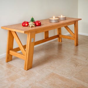 Puede incluir: Banco de madera marrón claro con una parte superior rectangular y patas en ángulo. El banco está decorado con manzanas rojas, un pequeño árbol de Navidad, una estrella dorada y dos velas plateadas encendidas. El banco mide unos 122 cm de largo.