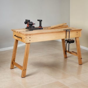 May include: A wooden workbench with a light brown finish. It features a black and orange lathe, a black vise, and a wooden plank. The workbench has a rectangular top and angled legs, suitable for woodworking projects.