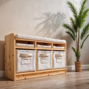 以下が含まれることがあります： 3つの布製ビンとクッション付きの天板が付いた木製の収納ベンチ。 ベンチは明るい色の木材でできており、シンプルでモダンなデザインです。 植木鉢がベンチの隣にあります。