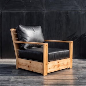 Könnte beinhalten: Ein moderner Outdoor-Sessel mit einem Rahmen aus Naturholz und schwarzen Lederkissen. Der Stuhl hat eine Lamellenlehne und Armlehnen und bietet eine Mischung aus Komfort und modernem Design. Der Stuhl steht vor einem dunklen Hintergrund.