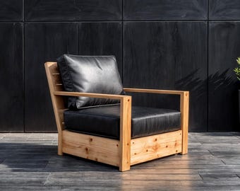 May include: A modern outdoor armchair with a natural wood frame and black leather cushions. The chair features a slatted back and armrests, offering a blend of comfort and contemporary design. The chair is set against a dark background.