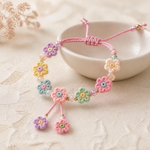 Può includere: Un braccialetto fatto a mano con ciondoli a forma di fiori all'uncinetto in colori pastello. Il braccialetto ha un cordino rosa con chiusura regolabile e piccole perline. I fiori sono rosa, gialli, verdi e viola.