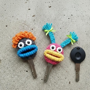 May include: Crocheted key covers in the shape of cartoon faces. One has orange hair and a blue mouth, the other has pink and yellow features with blue pigtails. A black car key is also shown.