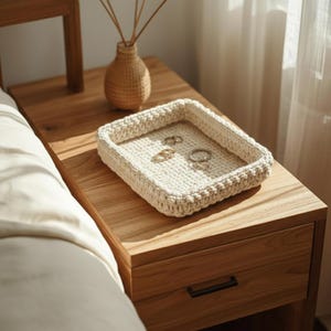May include: A rectangular, off-white, crocheted tray with a textured surface sits on a wooden nightstand. The tray holds several rings. A small, woven vase with dried stems is also on the nightstand.