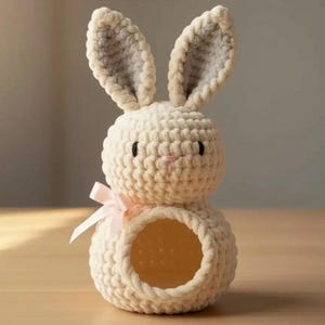 Könnte beinhalten: Ein cremefarbener, gehäkelter Hasenkorb mit einer rosa Schleife. Der Hase hat große, aufrechte Ohren mit grauem Innenfutter, schwarze Augen und eine rosa Nase. Der Korb hat eine kreisförmige Öffnung im unteren Bereich.